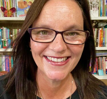 A close-up image of a white woman with long straight brown hair and glasses, smiling in front of a bookshelf