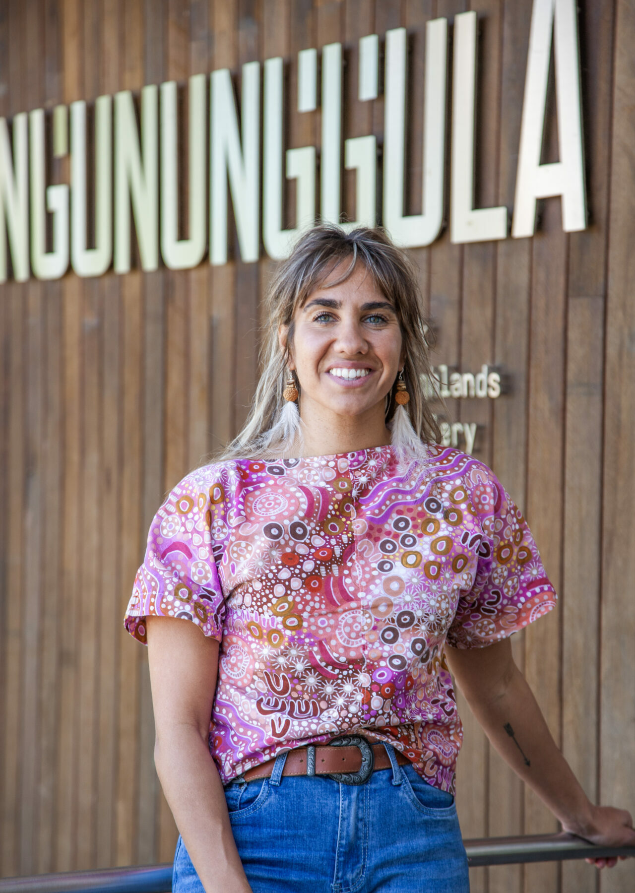 A brown-skinned woman with straight grey-brown hair smiles, leaning against a barrier, wearing a pink top patterned with Aboriginal art