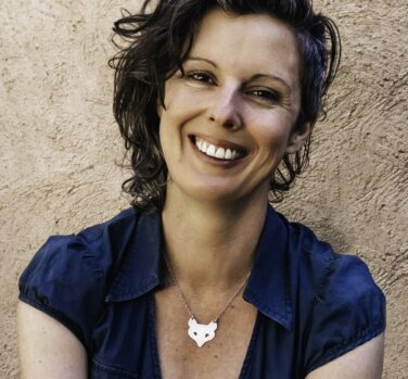 A woman with short wavy dark hair smiles widely at the camera, wearing a teal blouse