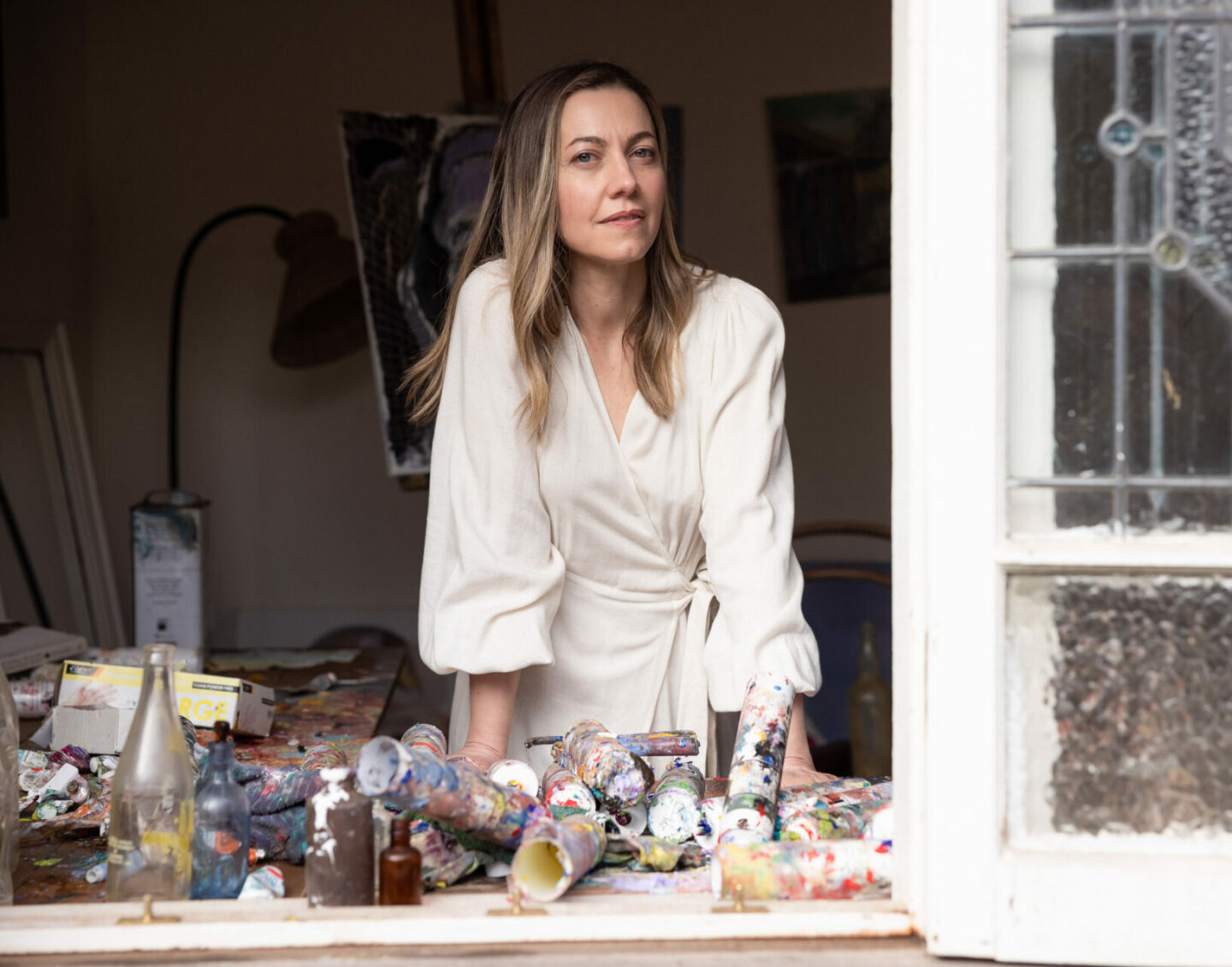 A white woman with long dark-blonde hair leans on a windowsill covered in art supplies
