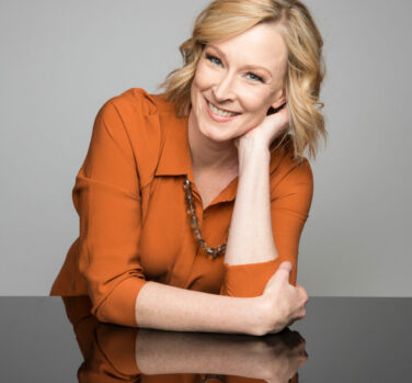 A middle-aged blonde woman leans on one hand, elbow resting on a desk, and smiles widely