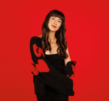 A brunette woman with a fringe stands against a red background, a sweater off one shoulder