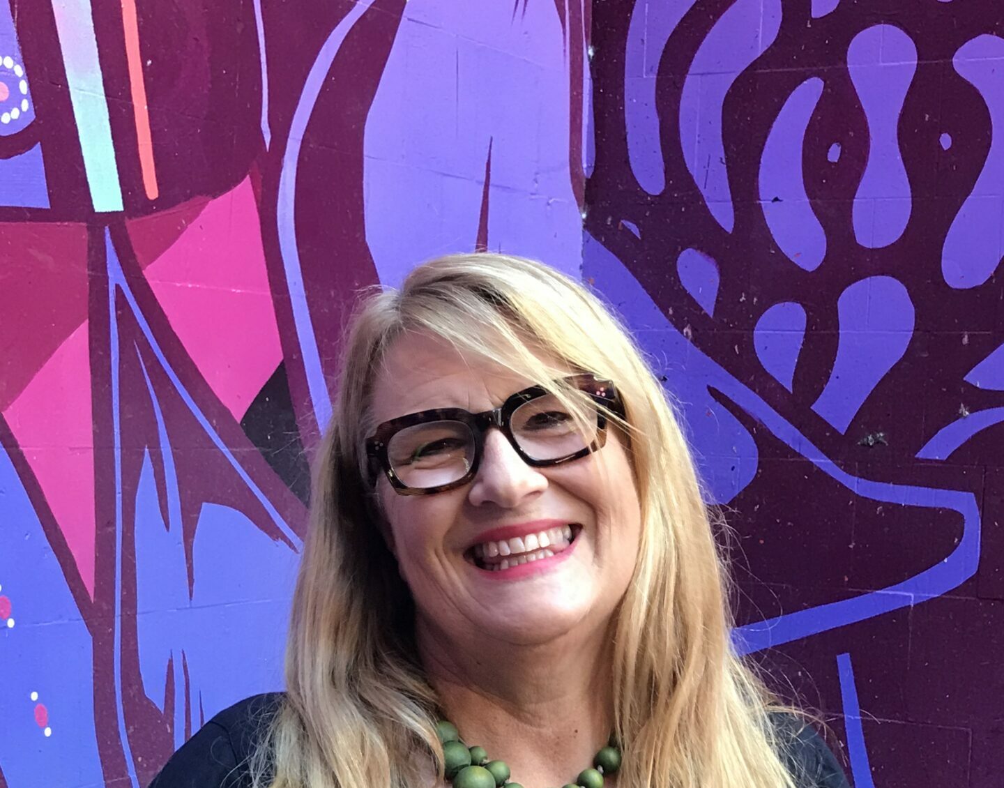 A middle-aged blonde woman with thick dark-rimmed glasses stands smiling in front of a blue and purple artwork