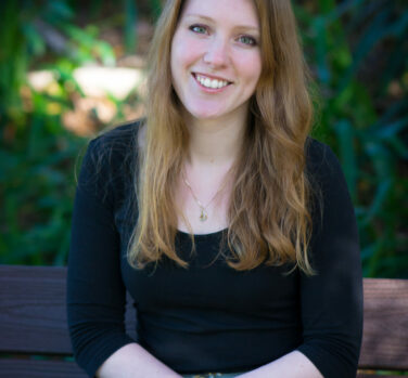 A young white woman with long light brown hair sits with her hands in her lap on a bench and smiles