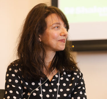 A white woman with shoulder-length brown hair looks off to one side and smiles. She is wearing and black and white spotted shirt