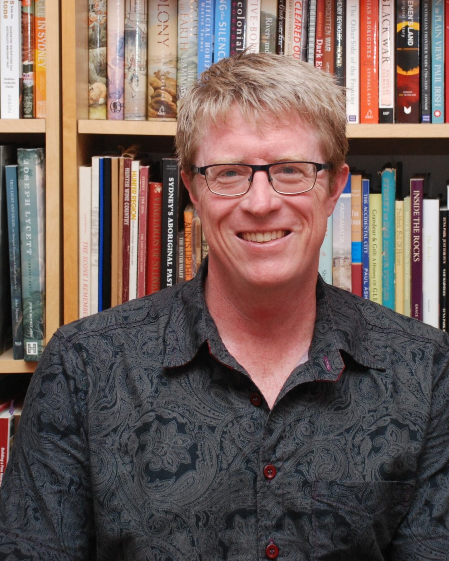 A blond man with dark-rimmed glasses and a black shirt smiles in front of a bookshelf