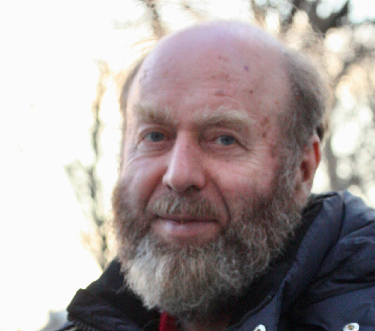 A photograph of a bald man with a bushy gray beard standing tilted away from the camera in a thick jacket