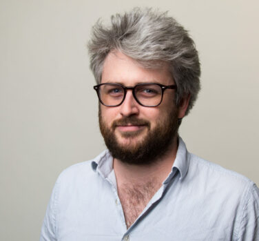 A white man with thick gray hair and a brown beard, wearing dark-rimmed glasses, smiles closed-lipped at the camera