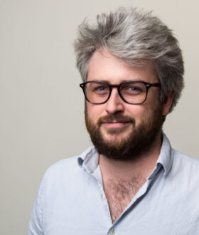 A white man with thick gray hair and a brown beard, wearing dark-rimmed glasses, smiles closed-lipped at the camera