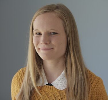 A young white woman with straight dark-blonde hair smiles closed-lipped at the camera, wearing a yellow jumper