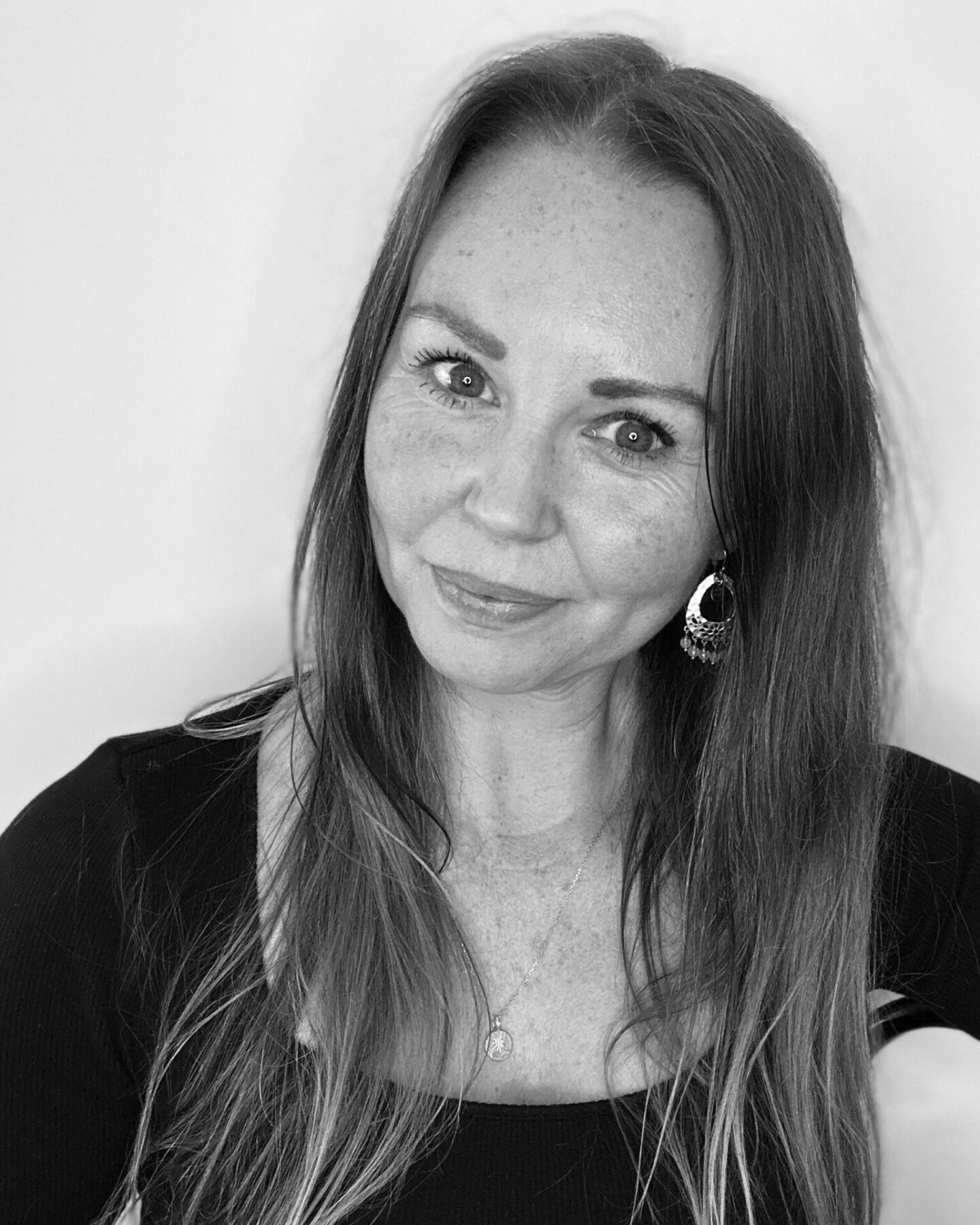 A black and white photograph of a woman with long straight hair and a black top, looking directly at the camera with her head tilted and a small smile