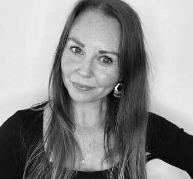 A black and white photograph of a woman with long straight hair and a black top, looking directly at the camera with her head tilted and a small smile