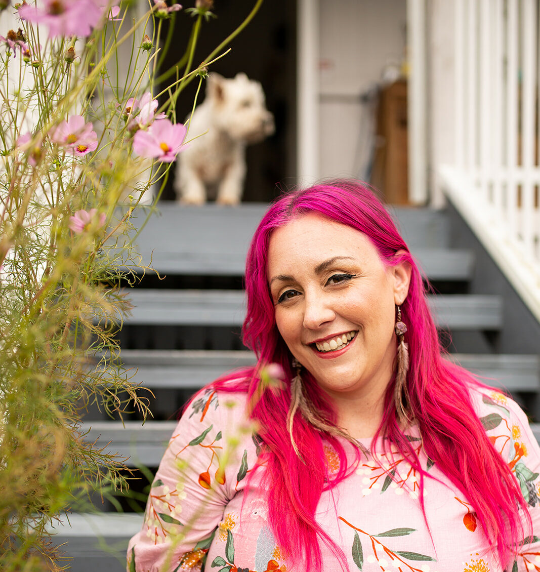 A woman with long bright-pink hair and a pale pink floral dress sits on some steps and smiles