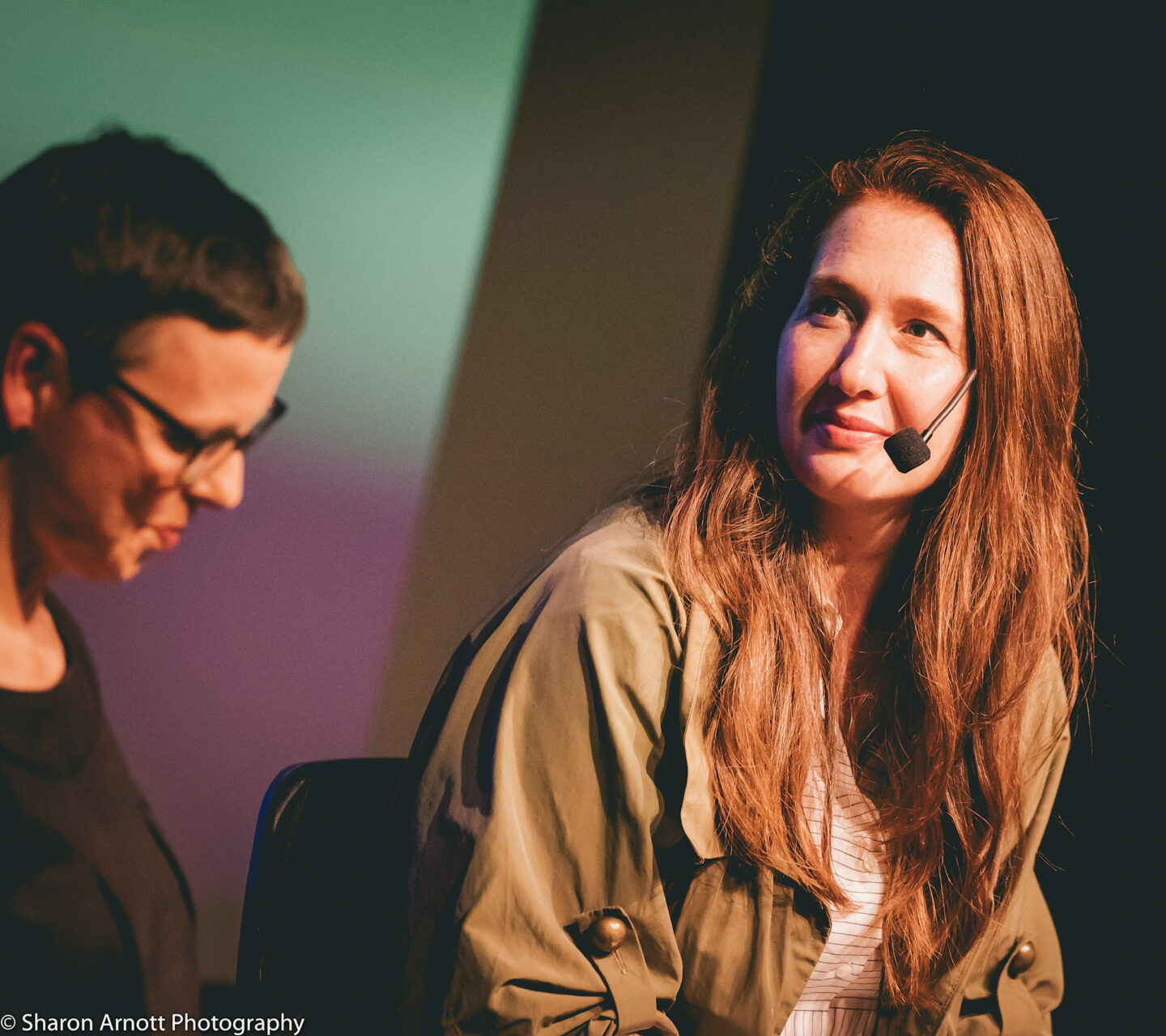 A woman with long brown hair wearing a head microphone is tilted side-on, talking to somebody