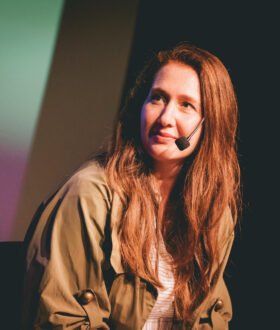 A woman with long brown hair wearing a head microphone is tilted side-on, talking to somebody