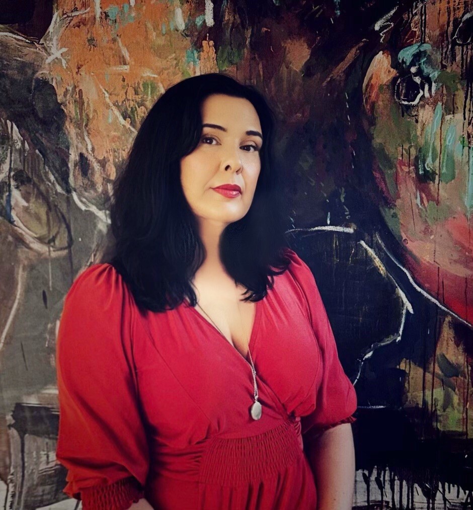 A woman with shoulder-length black hair, wearing red lipstick and a red dress, stands in front of a dark coloured artwork and looks at the camera without smiling