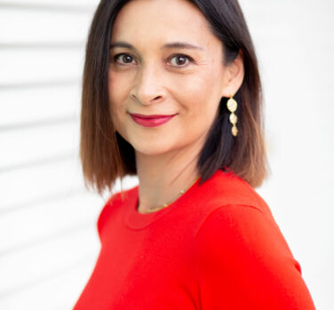 A woman with shoulder-length straight brown hair and a red top stands side-on, her head turned to smile at the camera