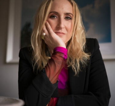 A blonde woman in a purple top an black blazer leans on one hand, sitting at a desk