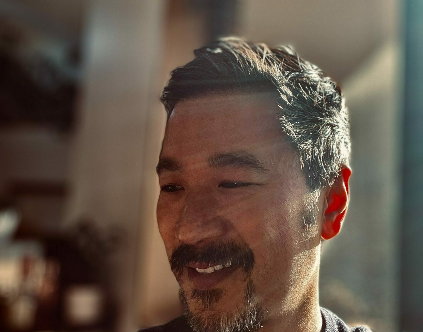 An Asian man with a goatee stands at an angle, smiling, as light shines across his face