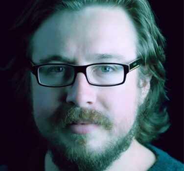 A close-up photograph of a young man with shoulder-length wavy hair, a medium-length brown beard, and rectangular black-framed glasses