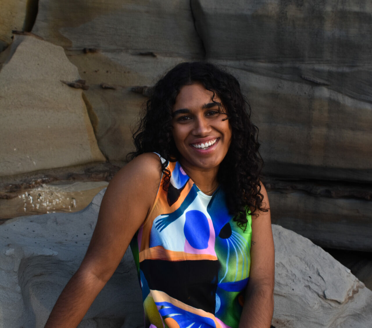 A dark-skinned woman with long curly black hair sits on a rock, smiling widely in a colourful dress