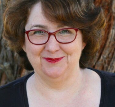 A middle-aged woman with short wavy brown hair, red glasses and bright lipstick smiles at the camera with closed lips