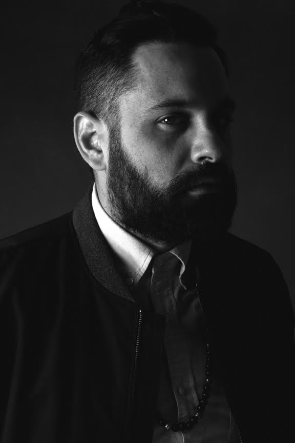 A black and white photograph of a bearded man in a white buttoned shirt and dark zippered jacket, half in shadow
