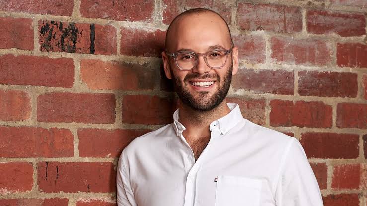 A bald man with a short brown beard and round thin-rimmed glasses smiles in front of a brick wall