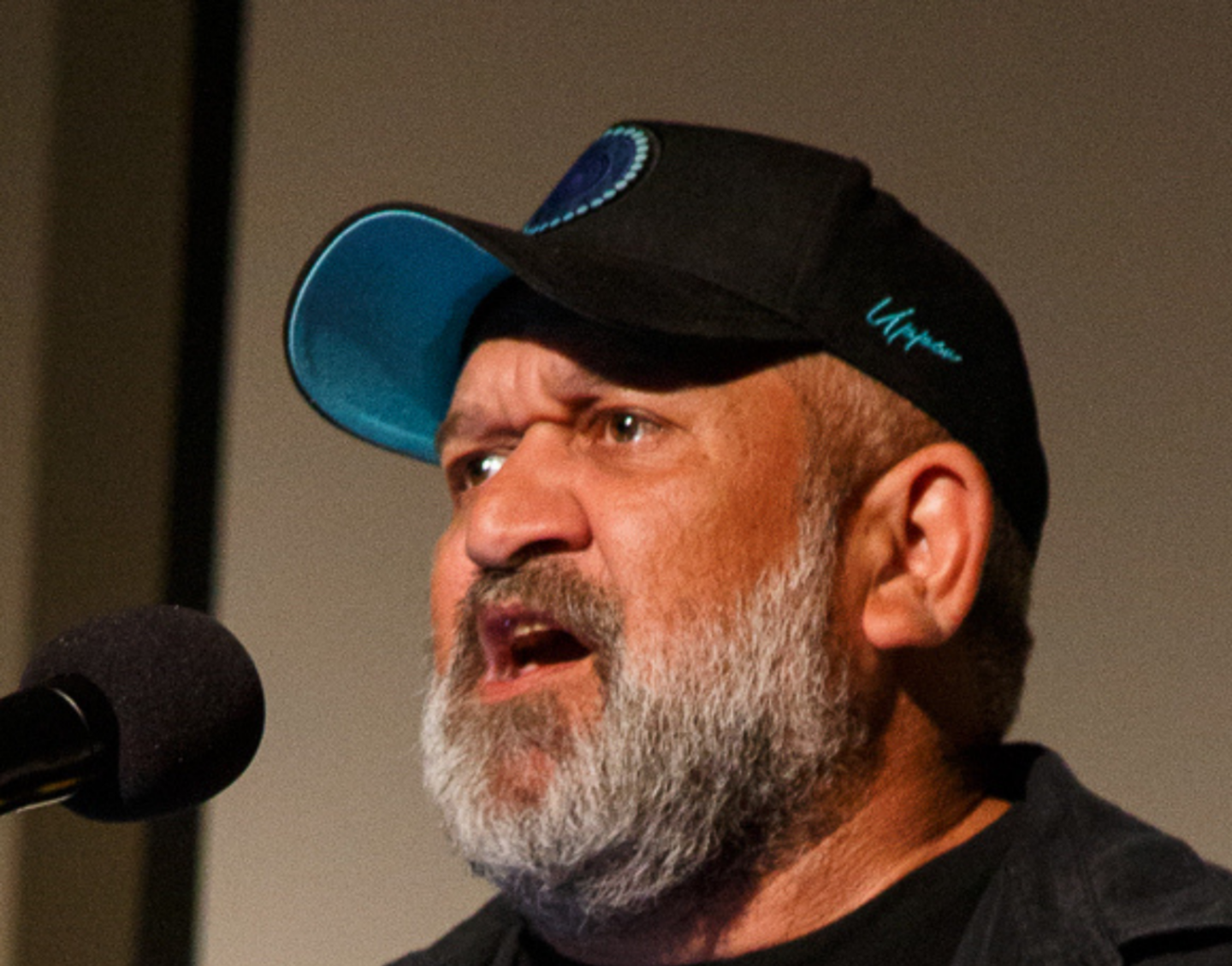 A man with a black cap and grey beard, wearing a shirt emblazoned with the Aboriginal flag. He is speaking into a microphone