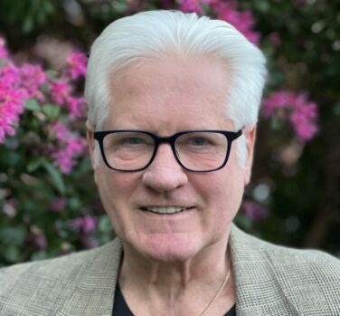 A man with short white hair and black glasses, wearing a grey blazer and smiling