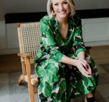 A blonde woman in a green patterned dress sits in a chair, leaning forward with her elbows on her knees and smiling