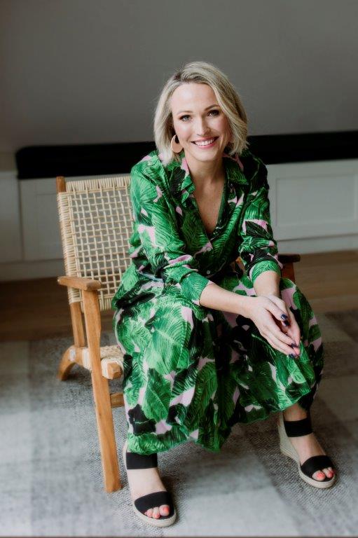 A blonde woman in a green patterned dress sits in a chair, leaning forward with her elbows on her knees and smiling