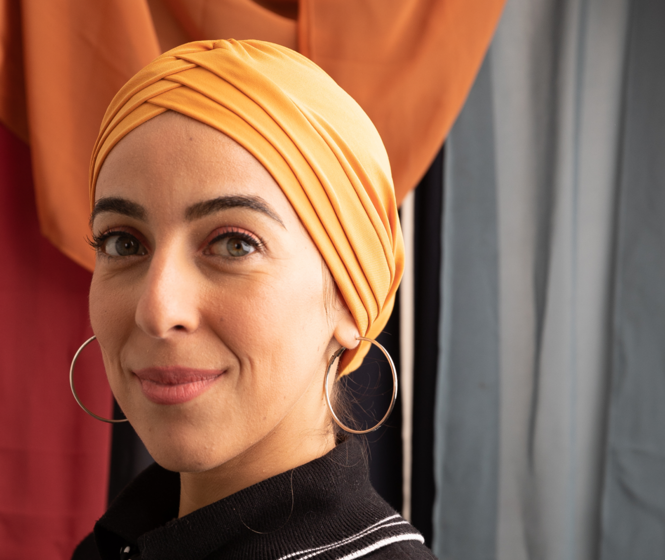 A woman with large hoop earrings and a yellow headscarf stands at an angle to the camera, smiling with closed lips