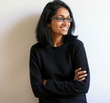 A dark-skinned woman with shoulder-length black hair and glasses stands arms folded, looking to one side and smiling widely