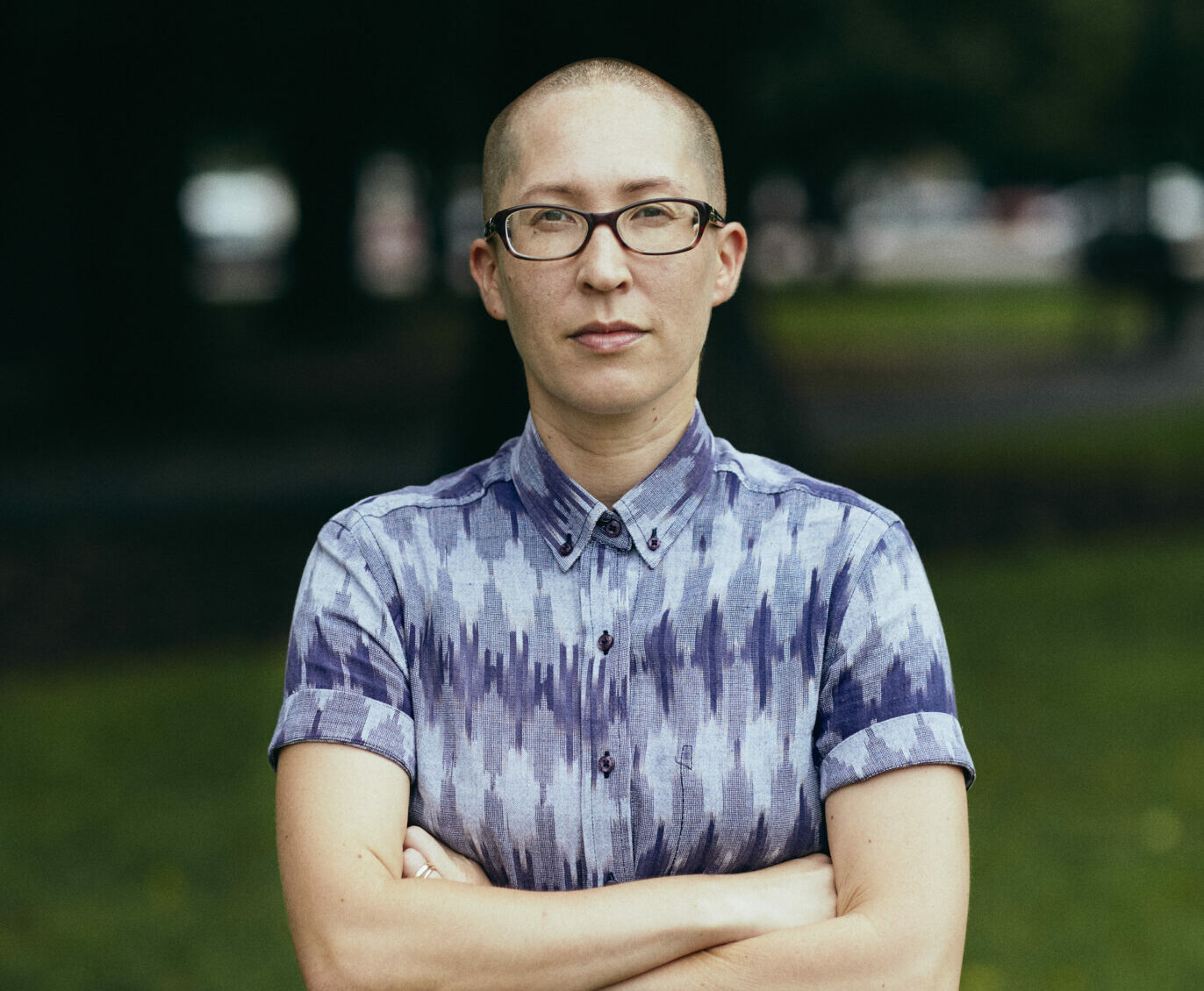 A person with a shaved head and dark-rimmed glasses stands arms-folded and looking directly at the camera