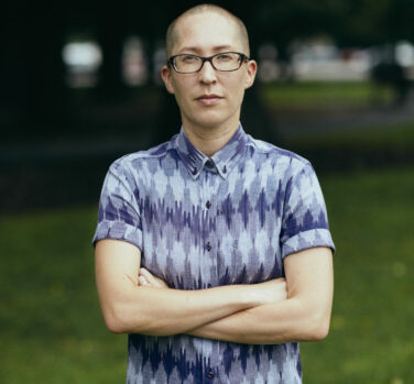 A person with a shaved head and dark-rimmed glasses stands arms-folded and looking directly at the camera