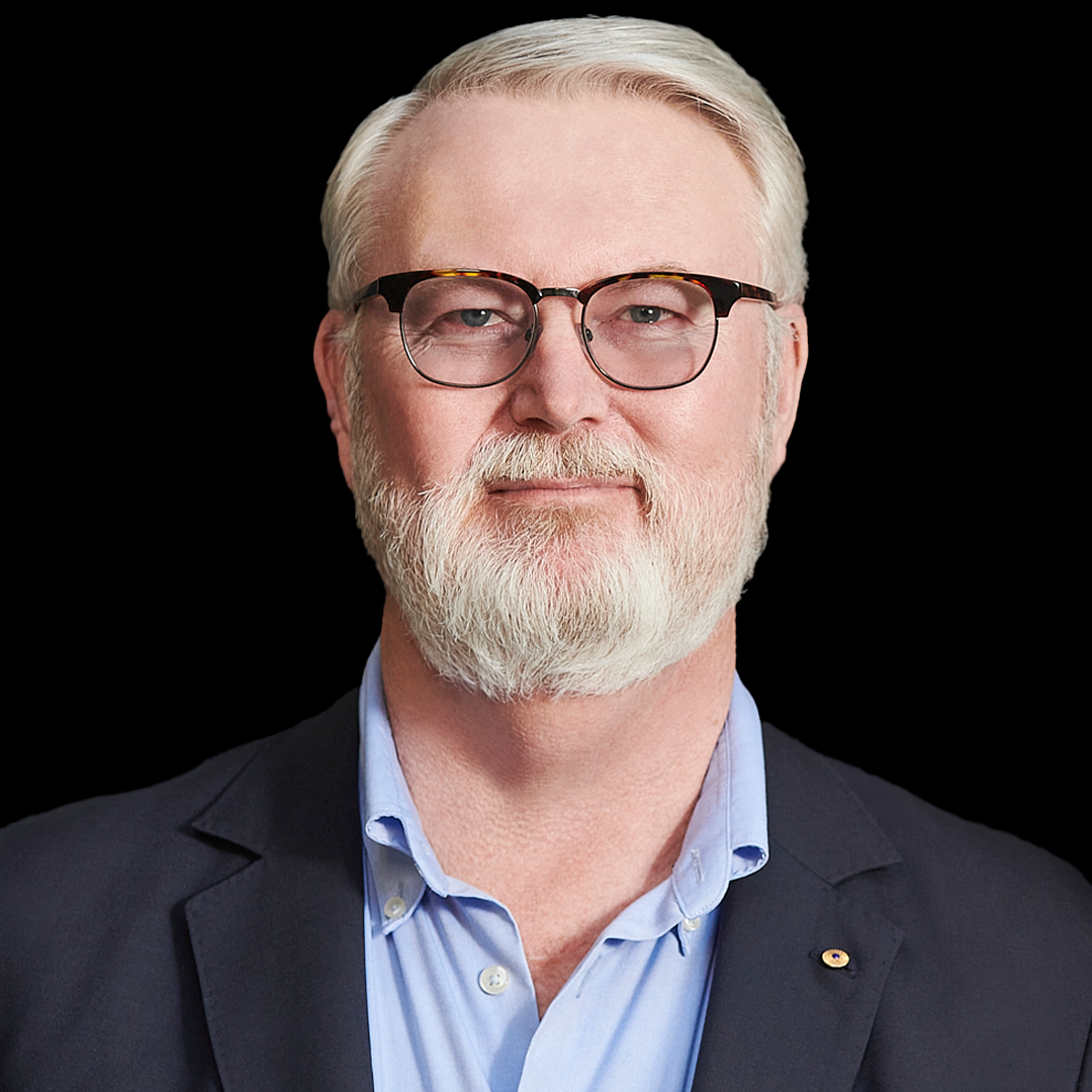 A man with white hair and full, trimmed beard, wearing dark-rimmed glasses and a dark gray blazer