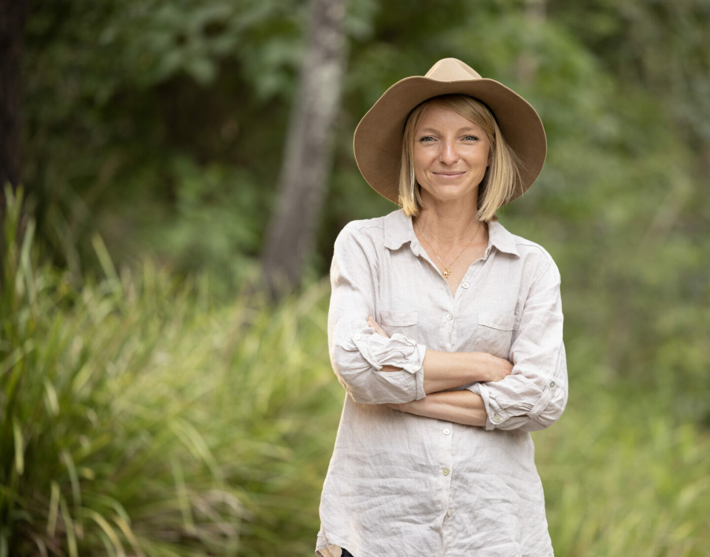 A blonde woman in a wide-brimmed hat stands outdoors, arms-folded and smiling