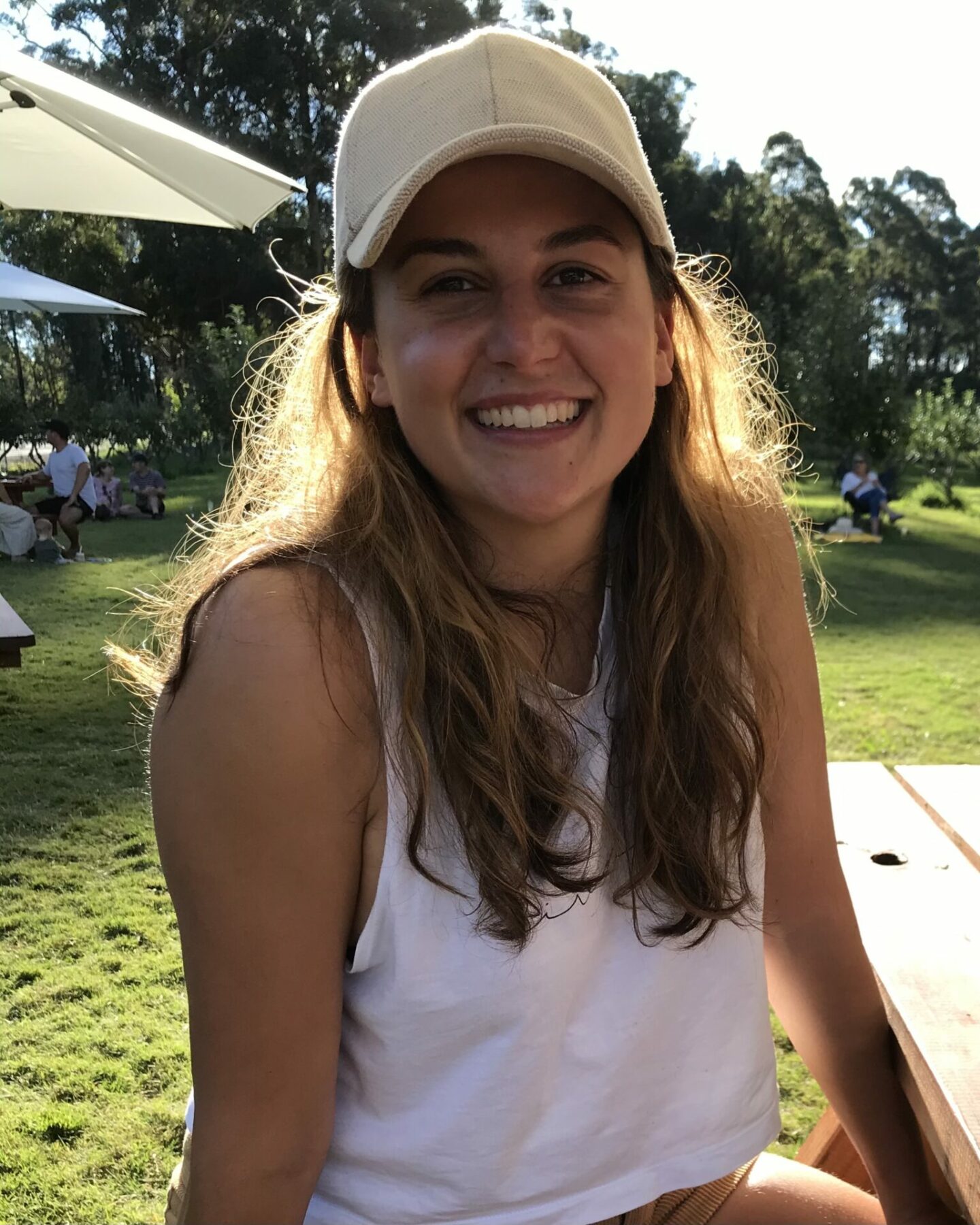 A young woman with long brown hair and a white cap smiles at the camera, sitting at a picnic table