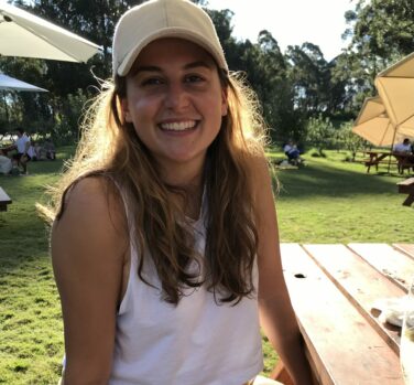 A young woman with long brown hair and a white cap smiles at the camera, sitting at a picnic table