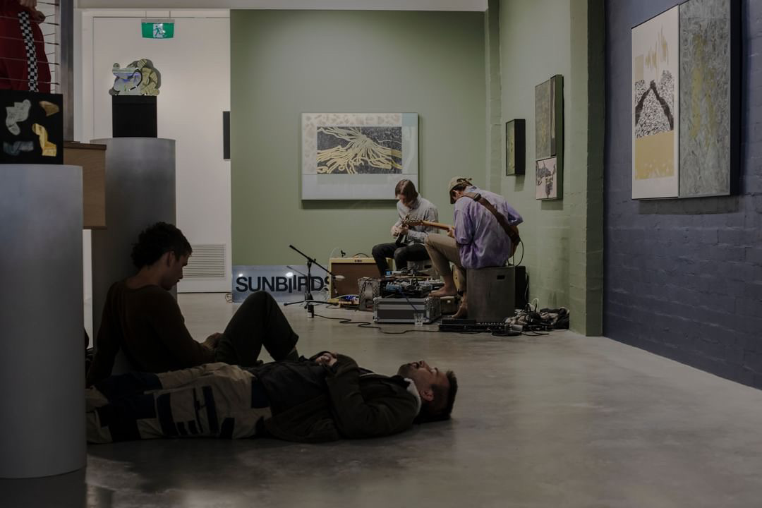 Four band members spread across a gallery room, one lying on the floor, one sitting beside him looking away from the camera, and two at the back looking down at their guitars