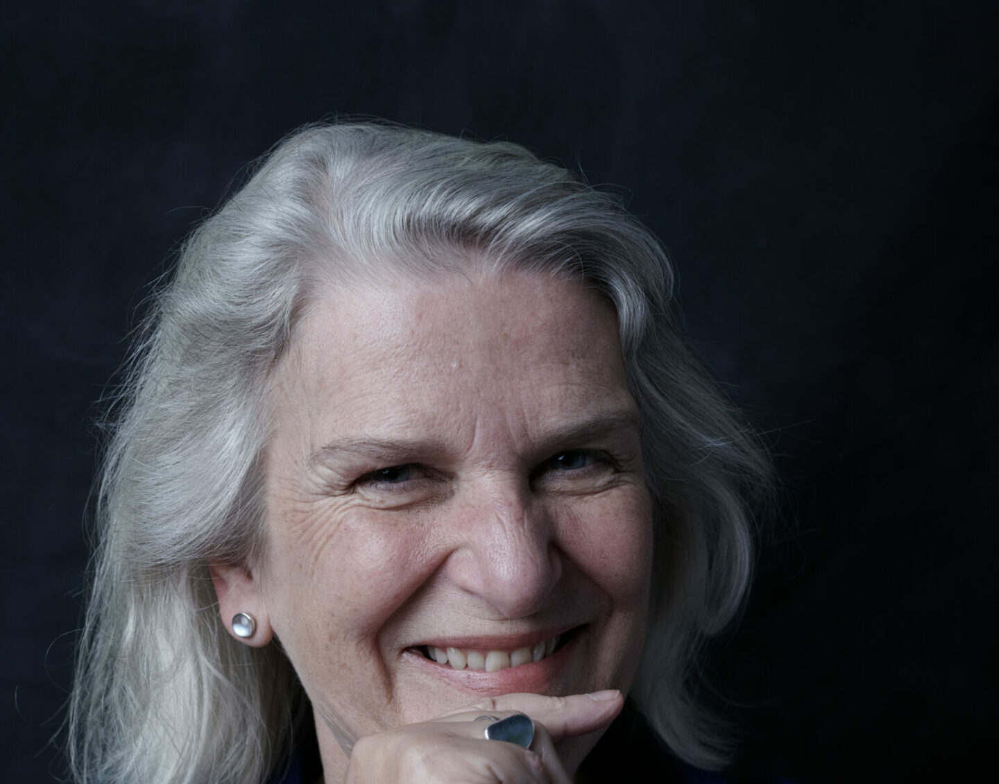 A woman with shoulder-length white hair smiles and rests her chin on a fist