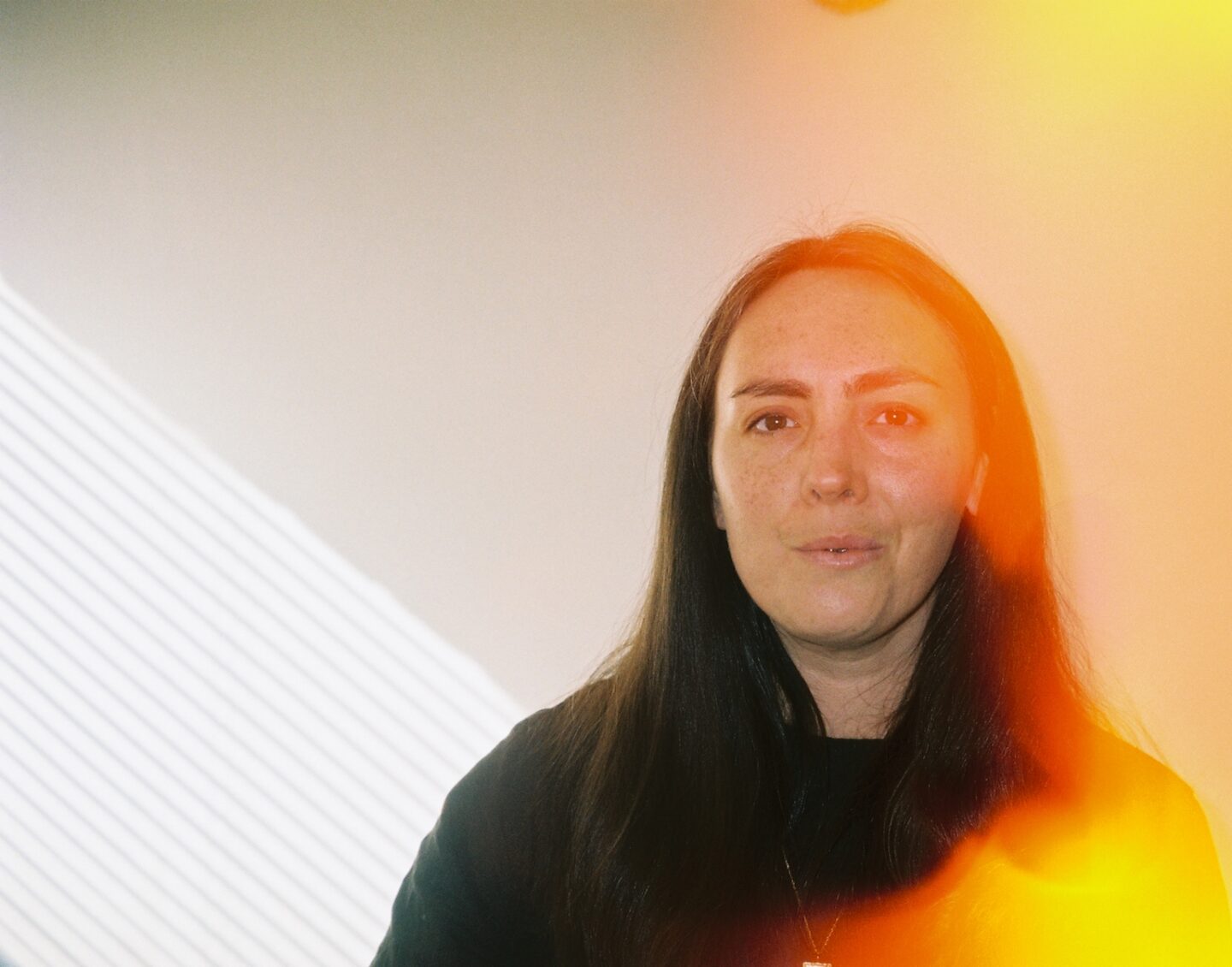 A middle-aged woman with long black hair looks directly at the camera, a yellow flare of light covering one side of her face and shoulder