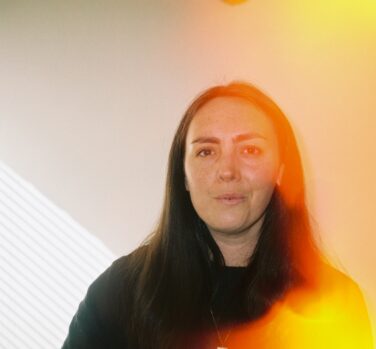 A middle-aged woman with long black hair looks directly at the camera, a yellow flare of light covering one side of her face and shoulder