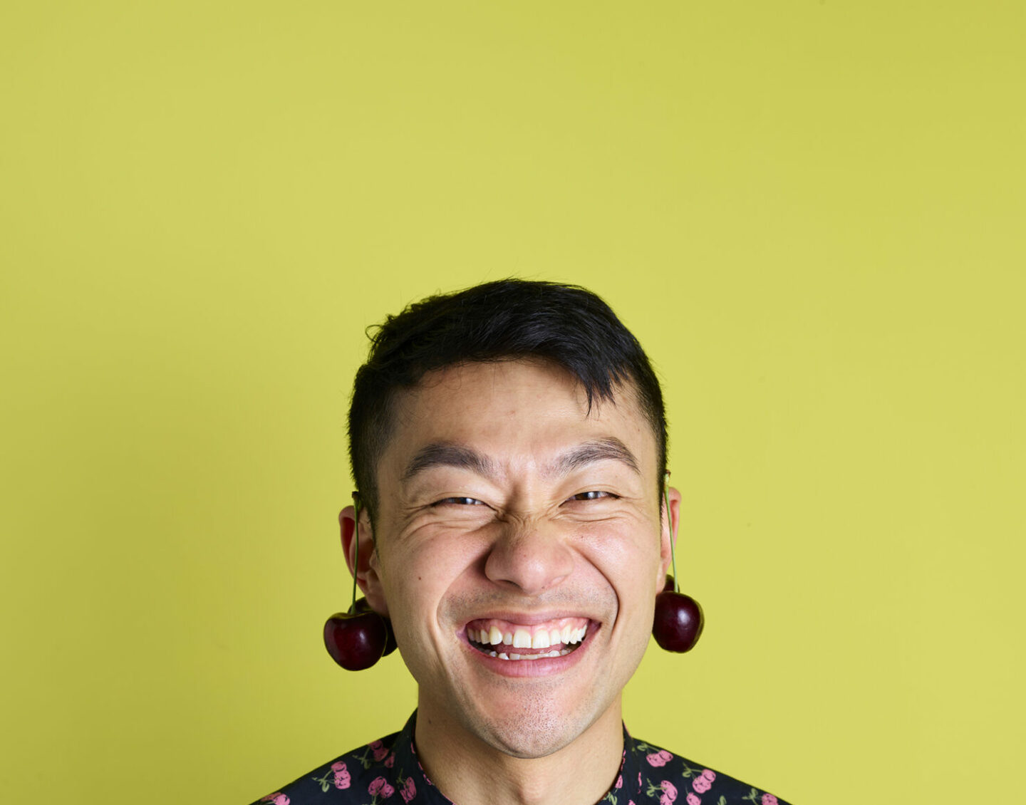 A black-haired man with cherries hanging over each ear grins at the camera in front of a yellow background