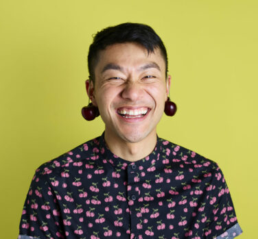 A black-haired man with cherries hanging over each ear grins at the camera in front of a yellow background