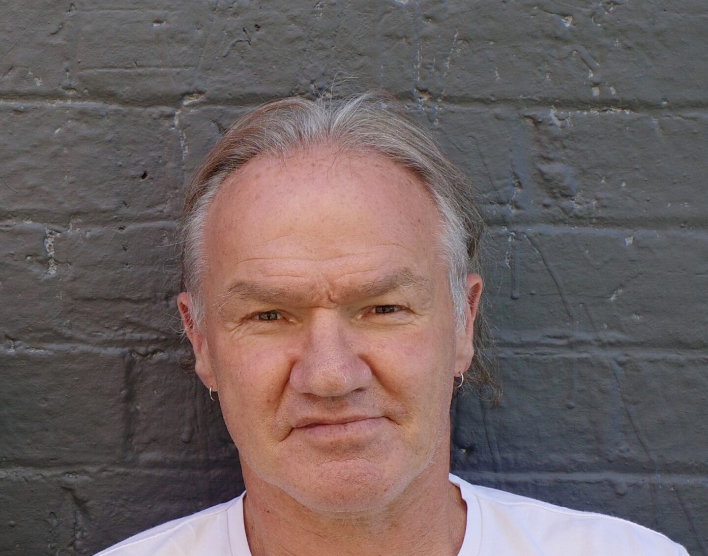 A man with short grey hair and a white tshirt stands in front of a grey painted brick wall