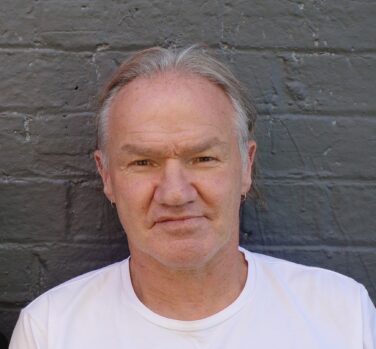 A man with short grey hair and a white tshirt stands in front of a grey painted brick wall