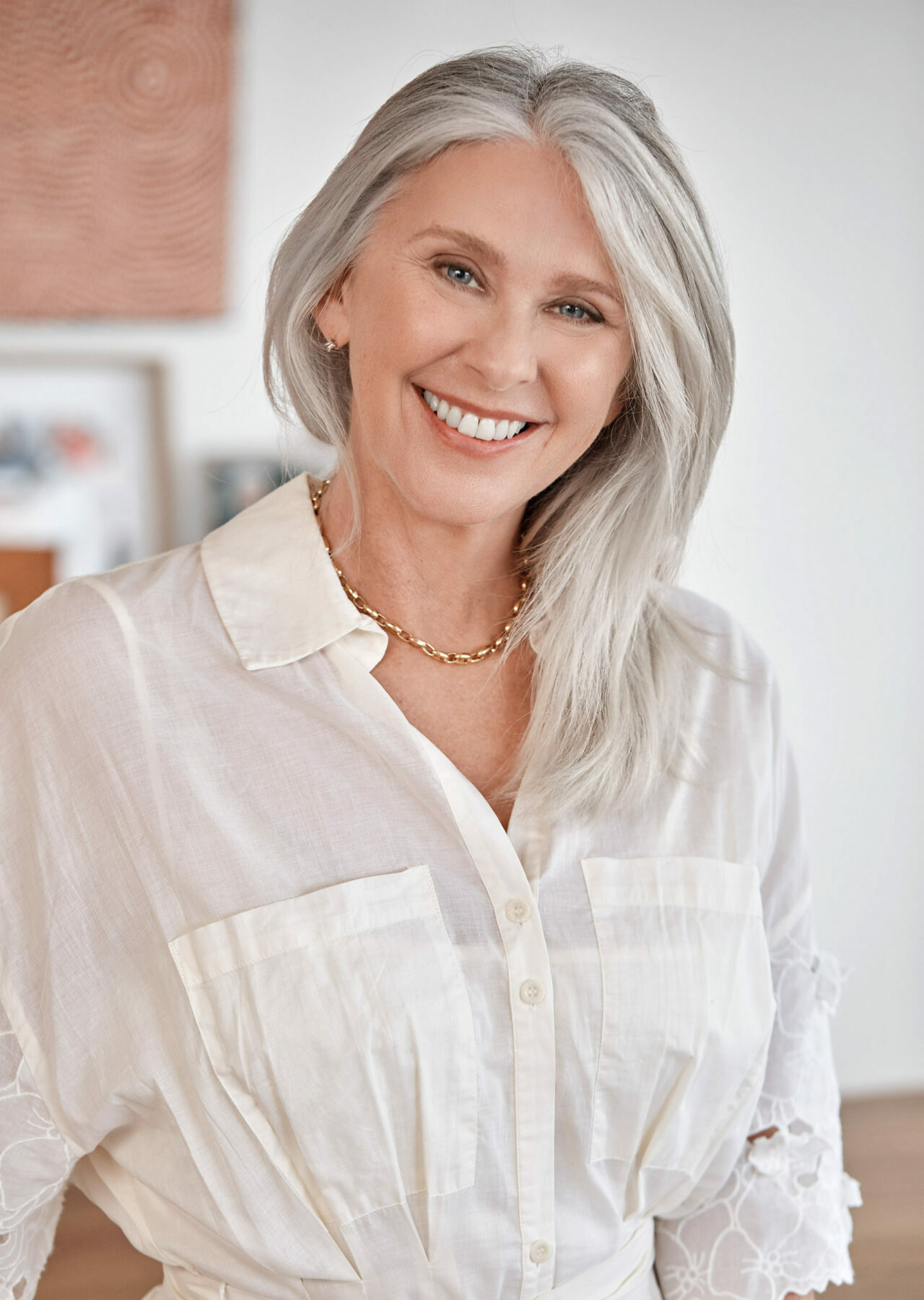 A woman with long grey hair and a button-up white dress smiles widely