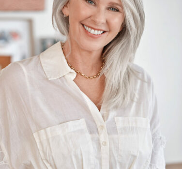 A woman with long grey hair and a button-up white dress smiles widely